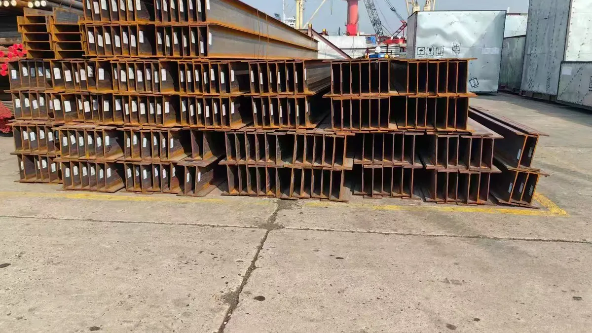 Yard stacks of H-beams showing end tags on webs against port crane background